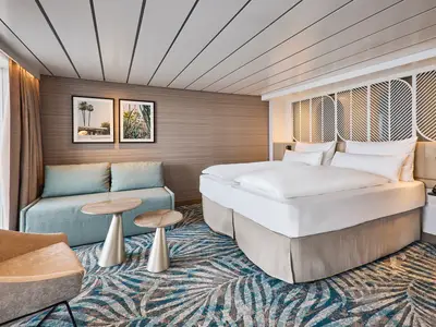 Helle Schöne Aussicht Suite an Bord der Mein Schiff Relax. Doppelbett, Sofa, Beistelltische und großes Fenster sind sichtbar. Tropische Kunst und Teppichmuster.