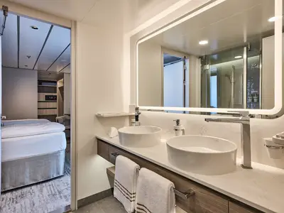 A brightly lit, modern bathroom on Mein Schiff Relax with a large mirror, two vessel sinks, and a walk-in shower reflected. The bedroom is visible through an open doorway.
