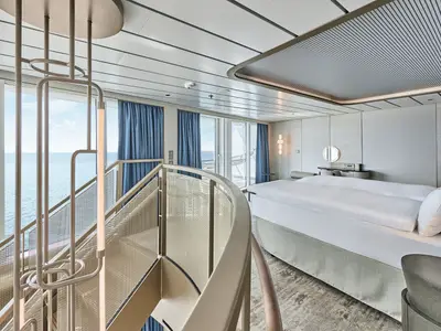Das helle Hauptschlafzimmer der Großen Freiheit Suite an Bord der Mein Schiff Relax. Ein großes Doppelbett, moderne Einrichtung, bodentiefe Fenster mit Meerblick und eine elegante Wendeltreppe sind zu sehen.