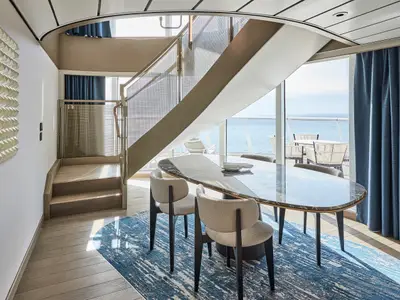 Moderner Essbereich einer Suite an Bord der Mein Schiff Relax. Ovaler Marmortisch, Stühle, blauer Teppich und eine elegante Treppe. Große Fenster bieten weiten Meerblick und zeigen einen Balkon.