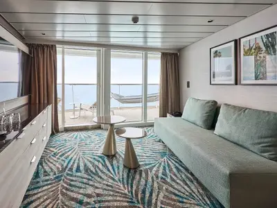 Moderner Wohnbereich der Fernweh Suite an Bord der Mein Schiff Relax. Hellblaues Sofa, zwei Tische und Balkon mit Meerblick.