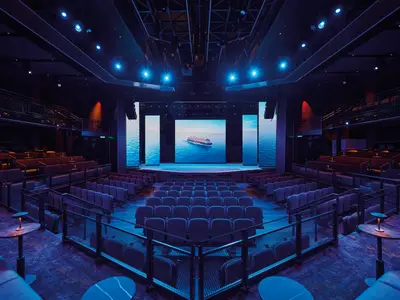 Das leere, moderne Theater an Bord der Mein Schiff Relax. Die große LED-Bühnenwand zeigt ein Schiff auf dem Meer. Blaue und orange Sitze auf Ebenen. Indirektes blaues Licht, kleine Tischlampen.
