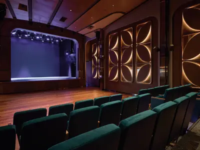 Eleganter Theatersaal der Mein Schiff Relax. Beleuchtete Bühne, teal-farbene Sitze, und moderne Holzpaneele mit Lichtinstallationen an den Wänden. Bereit für eine Show.